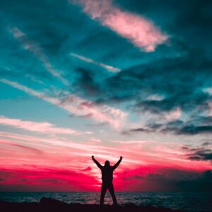 Person silhouetted against a vibrant sunset sky over the ocean, arms raised triumphantly.